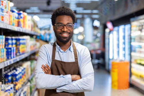 Obraz na plátne Young man wearing an apron and eyeglasses, smiling and looking at camera with ar