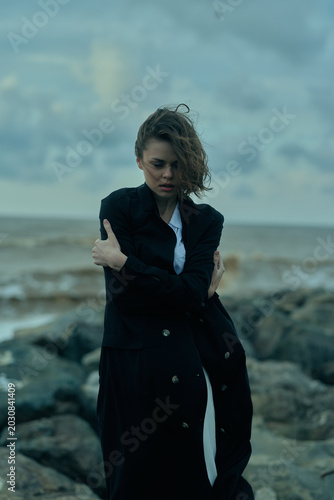 Bilde på lerret Woman in a long dark coat on a rocky shoreline, windblown hair, moody seascape highlighting fashion and resilience, dramatic palette