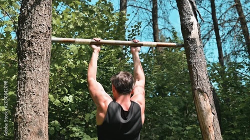 A young man performs pull-ups on a wooden bar set between two trees in a forest. The activity showcases strength training in a natural setting during daylight hours
