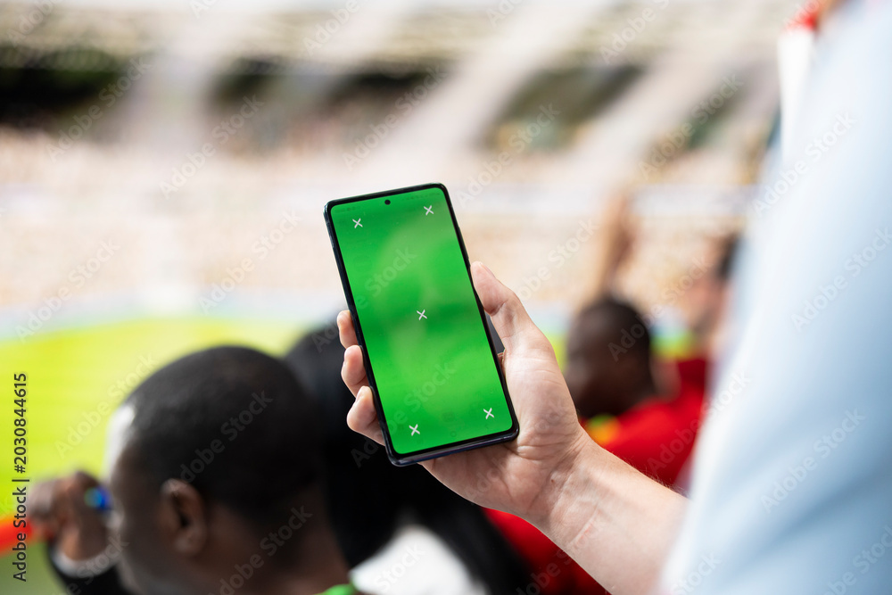 Obraz premium Excited football fans gather near a green screen phone mockup in stadium tribune for screen replacement and compositing.
