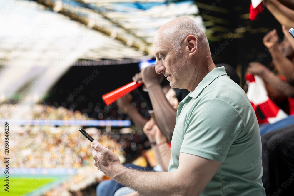 Obraz premium Focused football fan placing live bet ticket through mobile gambling app during soccer match with cheering supporters at the stadium. Man experiencing high risk betting tension with phone.