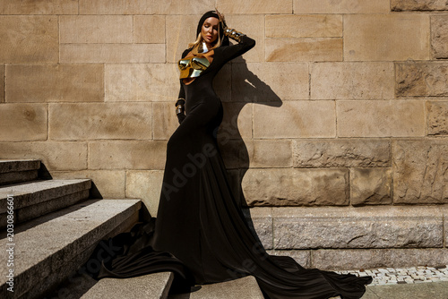 Foto Majestic female model in avant-garde flowing black gown with metallic gold armor and elaborate feather headdress posing against a textured stone wall in a high-fashion fantasy editorial