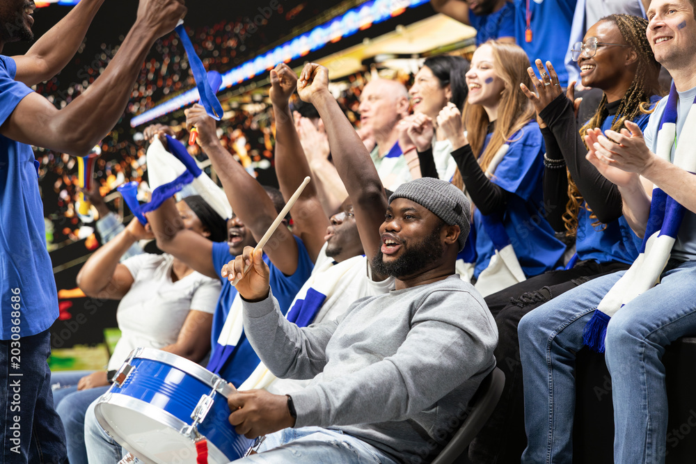 Obraz premium Black football supporter with drum leading blue stadium crowd chants while fans cheer around him during soccer match. Diverse soccer ultras express team spirit through synchronized chants.