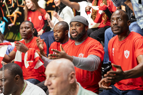 Schilderij op canvas Diverse sports fans reacting with suspense during football match in stadium stands, with red shirts and focused expressions