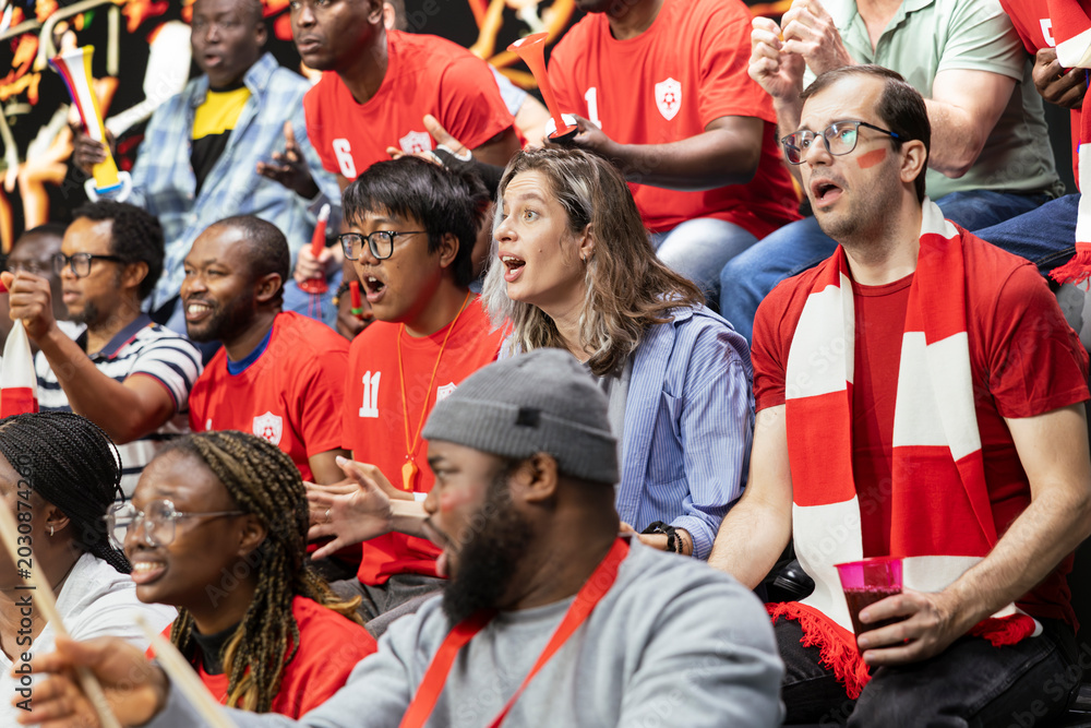 Fototapeta premium Red team soccer supporters watching tense football match from crowded stadium stands, focused faces, scarves and fan horns. Anxious diverse fans feeling game pressure after close call.