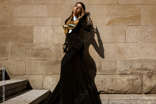 Fotografie Majestic female model in avant-garde flowing black gown with metallic gold armor and elaborate feather headdress posing against a textured stone wall in a high-fashion fantasy editorial