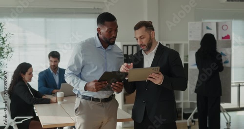 Portrait of African American and Middle Eastern businessmen check tablet in office as team works near desk. Colleagues compare data and discuss project
