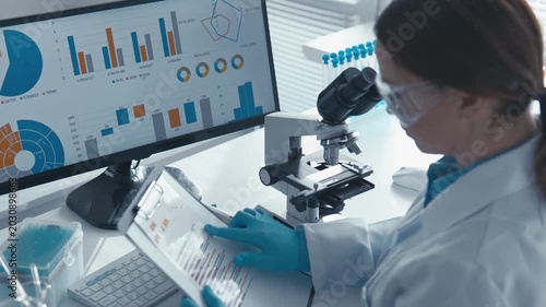 Woman researcher works in high tech lab, examining samples under microscope, reviewing analytics on tablet. Digital charts, laboratory equipment highlight scientific progress, innovation