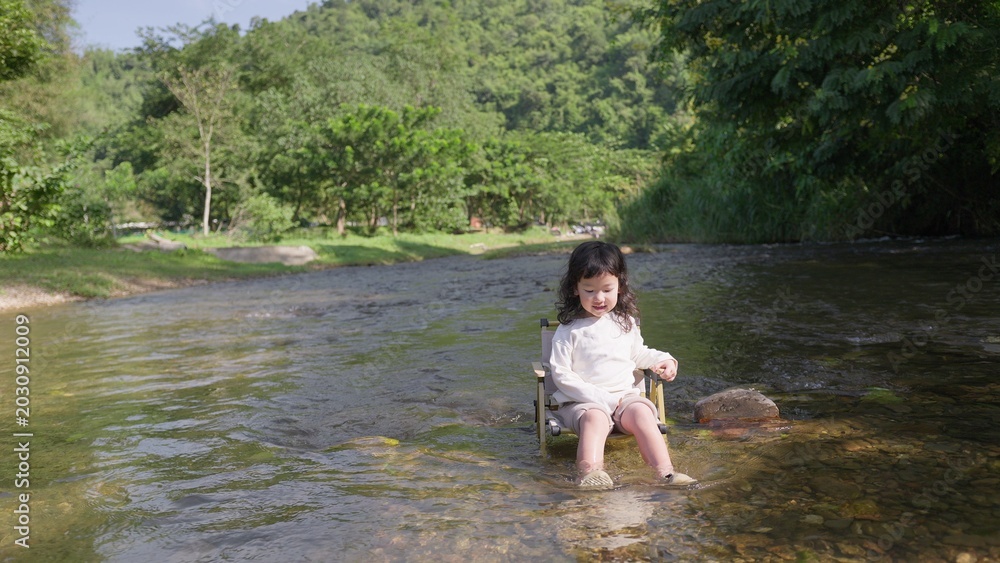 Fototapeta premium A young child sits peacefully on a camping chair in a shallow river, enjoying a quiet moment of sensory play and connecting with the natural environment.