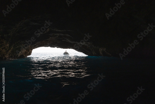 A small boat approaches the bright opening of a sea cave grotto, forming a dr...