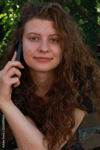 Young woman talks on phone while sitting in a garden during daylight hours with foliage in the background