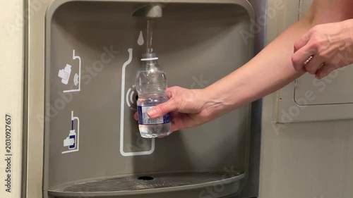 A person fills a plastic water bottle at a water fountain located in a school hallway while other students wait nearby. This happens during lunchtime.