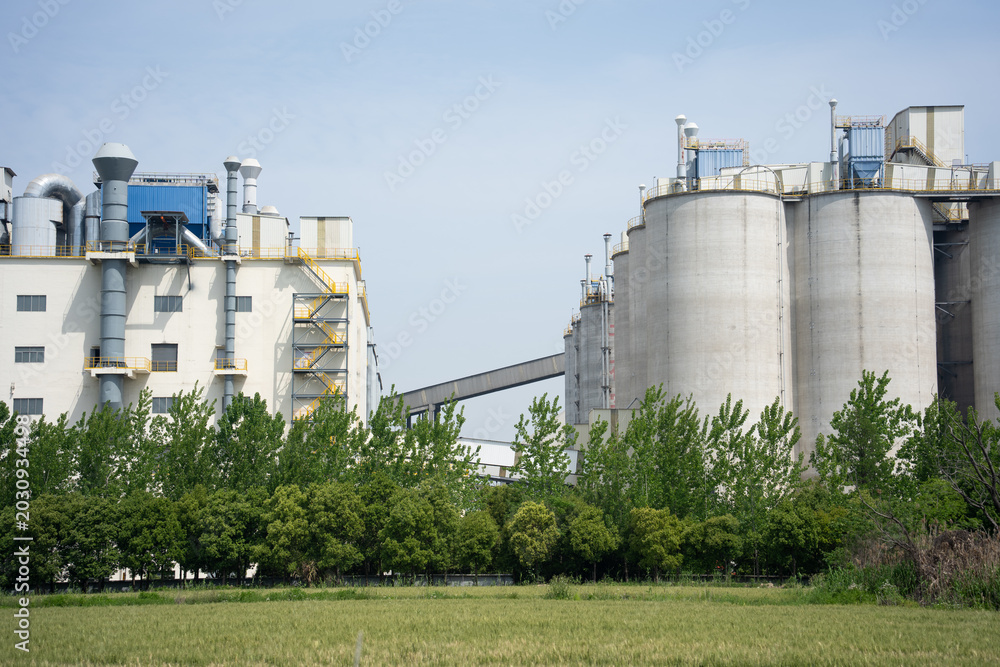 Fototapeta premium Modern industrial factory storage tanks hidden behind green trees under blue sky