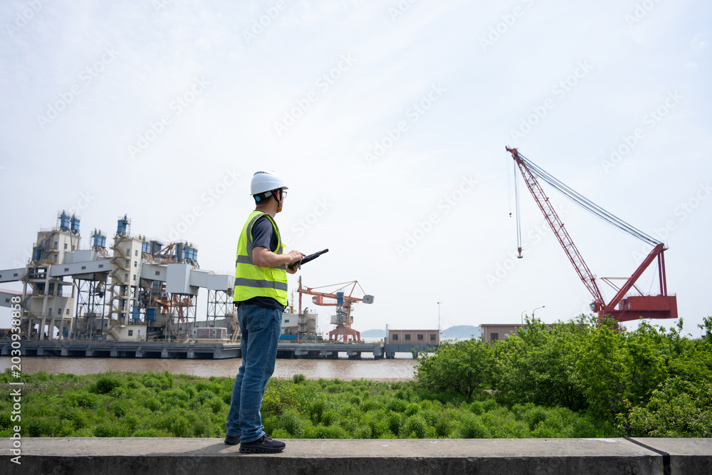 Fototapeta premium Engineer standing at port terminal holding tablet computer with cranes and industrial machinery in background