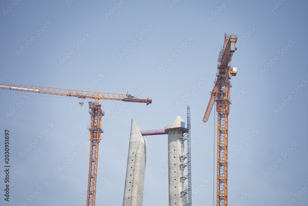 Fototapeta premium Two concrete bridge towers and yellow cranes against a clear blue sky background