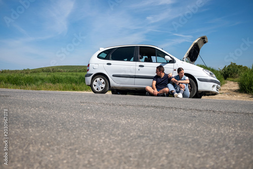 Canvas Print Couple sitting on asphalt road next to broken white car with open hood, showing