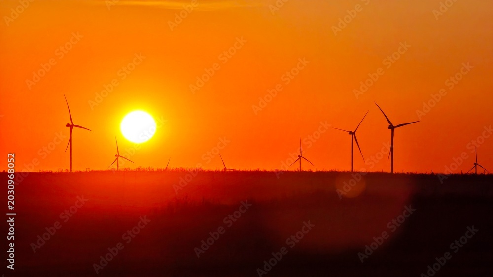 Fototapeta premium A vibrant sunset over a landscape with several wind turbines silhouetted against the orange sky. The scene captures renewable energy and natural beauty.