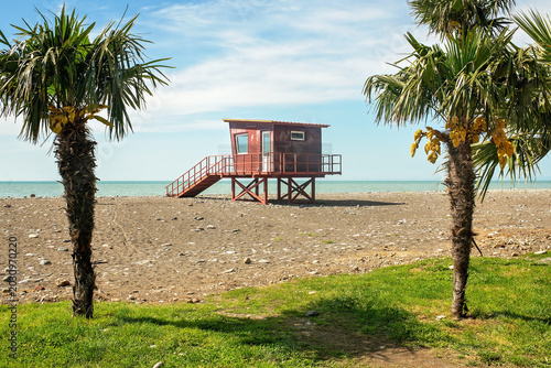Foto A red wooden lifeguard tower on a pebbly beach, enclosed with safety netting during the off-season, framed by palm trees against a blue sky