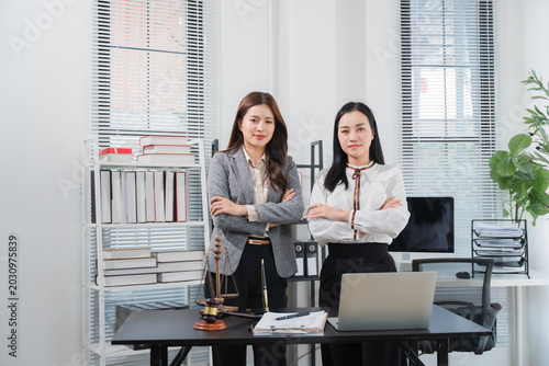 Lawyer Partnership in Office: Two confident female lawyers stand with arms crossed in a modern office setting, surrounded by legal books, a laptop, and scales of justice.