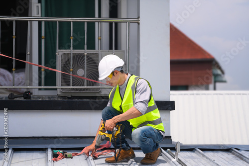 Fotografie Asian Male Roofer Using Power Drill on Metal Roof Installation with Safety Equip