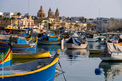 Tableau sur toile Traditional Maltese fishing boats moored in Marsaxlokk harbor with the Parish Church of Our Lady of Pompei in background, Malta