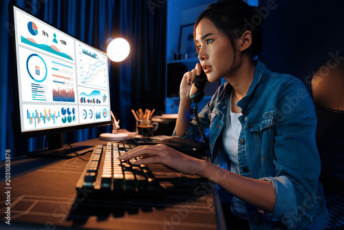 Konstfotografi Young beautiful Asian creative woman calling on phone to customer or coworker informing strategy marketing creator plan while searching market analysis on pc at neon modern office at night