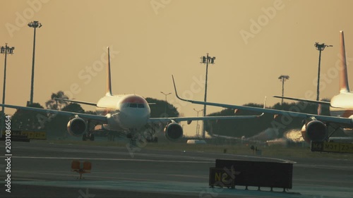 Airplanes taxi on runway at sunset in busy airport with tall lights and distant structures