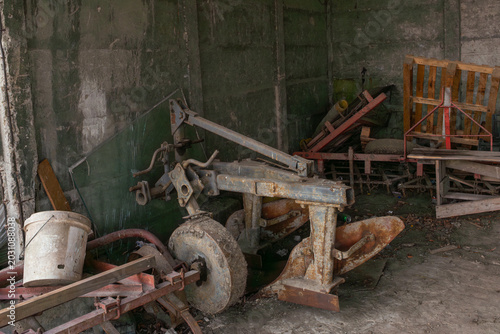 Photos Rusted plow and old agricultural implements stored in a dark concrete shed with pallets, debris, tire, and worn machinery parts