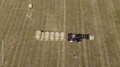 Tractor Harvesting Hay Bales in Field: An aerial view showcases a tractor actively collecting and transporting round hay bales across a dry, vast field under a clear sky.