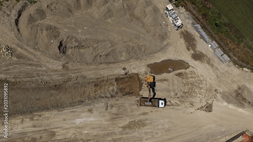 An excavator operates in a large sand quarry, with a cement truck visible in the distance, showcasing heavy machinery at work in an industrial setting.
