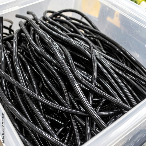 A clear plastic bulk bin full of black licorice whips in bright store lighting. Ai