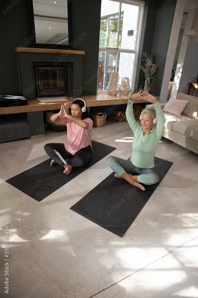Fototapeta premium Two mature women sitting on black yoga mats in living room near fireplace, one wearing headphones