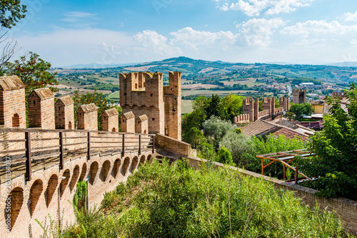 Ancient Gradara castle walls standing tall under a clear sky, featuring crenellated towers and arched structures, overlooking rolling hills and a picturesque Italian landscape in the Marches region