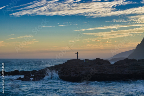 Schilderij op canvas A solitary angler stands silhouetted against the dawn sky, holding a fishing rod