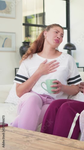 Vertical video: Smiling woman white tee taking hand as friend talking on couch with green pink mugs