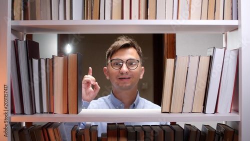 Tableau sur toile Young man wearing eyeglasses standing between bookshelves, having an exciting eu