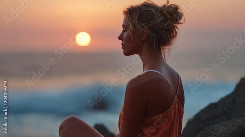 Young woman sitting on a rocky cliff overlooking the ocean at sunset. she is wearing a sleeveless orange tank top and her hair is styled in a messy bun.