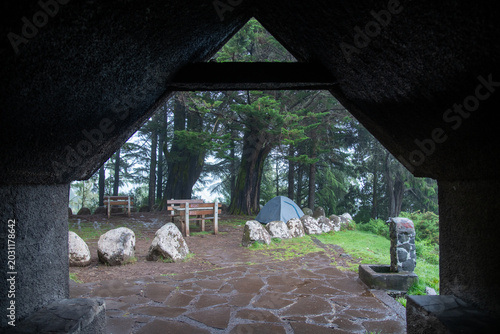 Cuadro en lienzo View from a stone shelter looking out at a green forest campsite with a gray tent and wooden picnic tables