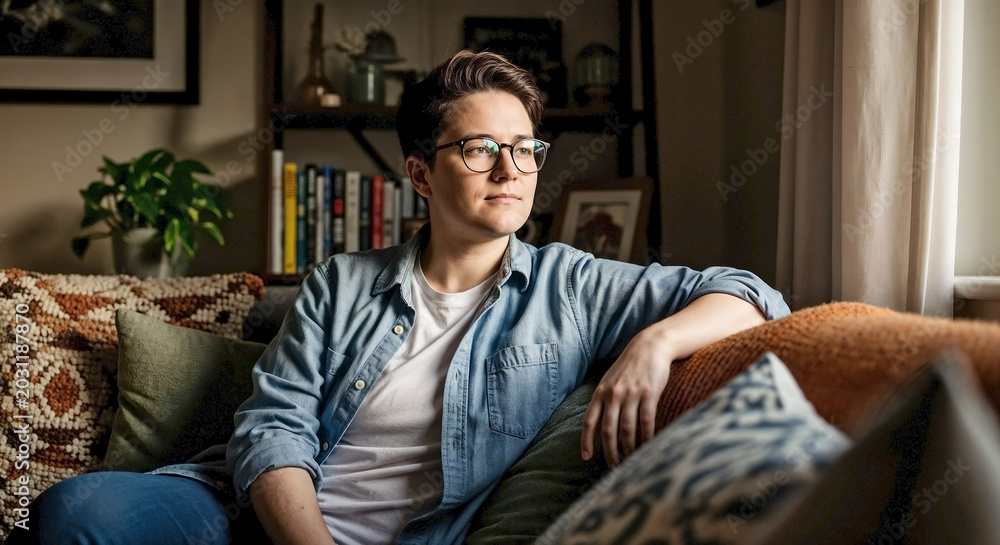 Obraz premium Non-binary person relaxing on home sofa, looking calmly out window. Non-binary person wears glasses, denim shirt, sitting comfortably amidst bright living room decor.