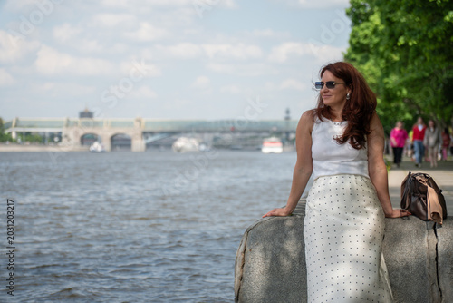 Photography Red-haired woman in sunglasses sits on Gorky Park embankment beside Moscow River with green riverside background
