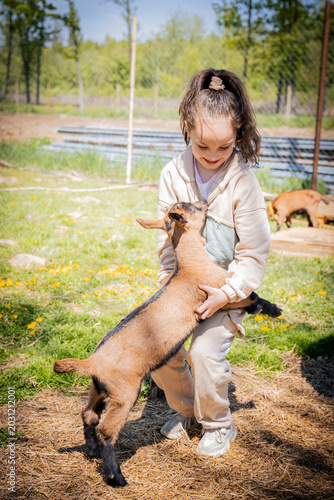 A young girl joyfully interacts with a playful goat on a sunny day, surrounded by greenery and farm animals.