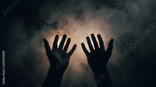 Fotografie Two Hands Reaching Upwards Towards Light and Smoke in Darkness