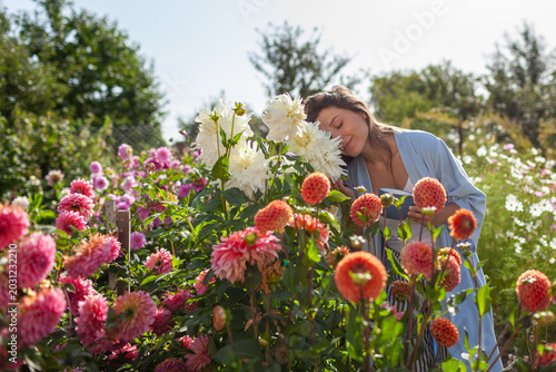 Woman enjoys blooming dahlias in summer garden drinking morning coffee in pajamas. Gardener walks by rows of flowers wearing robe