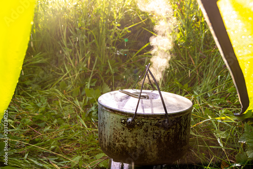 Old metal camping pot boiling on portable stove under yellow tent with steam rising and rain drops visible in backlight