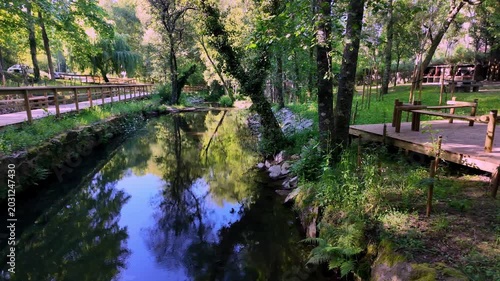 A peaceful river winds through a lush green park featuring wooden walkways and picnic platforms. The calm water mirrors the vibrant trees and blue sky during golden hour lighting.