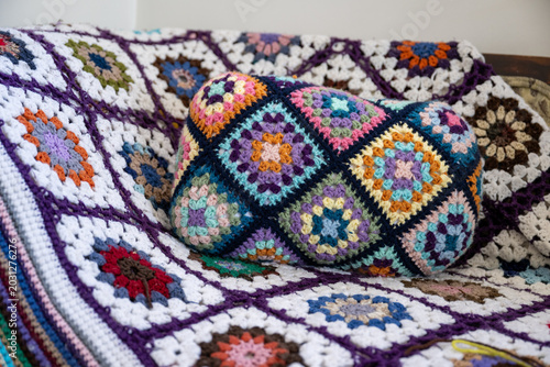 A colourful knitted blanket, handcrafted using interlocking loops of yarn, known as a common granny square crocheted blanket. There's a darker version of the pattern in the middle as a pillow. 