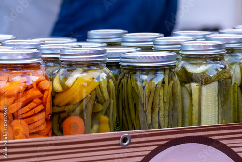 Glass jars of preserved pickles, beans and carrots soaking in garlic brine and spices on a tray. The vegetable-filled bottles are for sale at a farmers' market. The crocks are clear glass with lids.