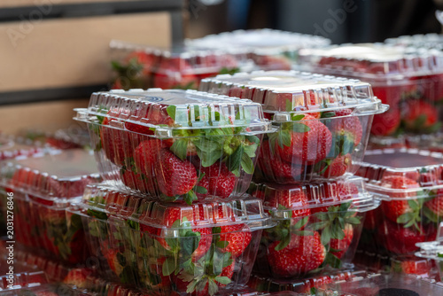 Clear plastic containers of fresh red, ripe strawberries with the stems still on them. The packages are stacked on top of each other on a grocery store shelf.  The berries are firm, rich, and vibrant.