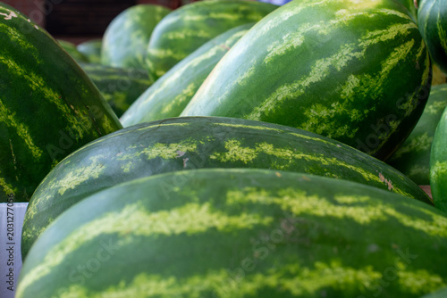 A stack of large, organic, oblong-shaped green and yellow striped watermelons on a table at a farmers' market. The African melons have thick peels with a striped pattern and a juicy interior. 