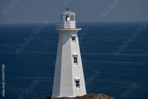 A white colored vintage, octagon-shaped lighthouse, lantern room, and tower containing storm glass panes. There's a rocky coastline in the foreground. The background is a blue ocean and a blue sky.